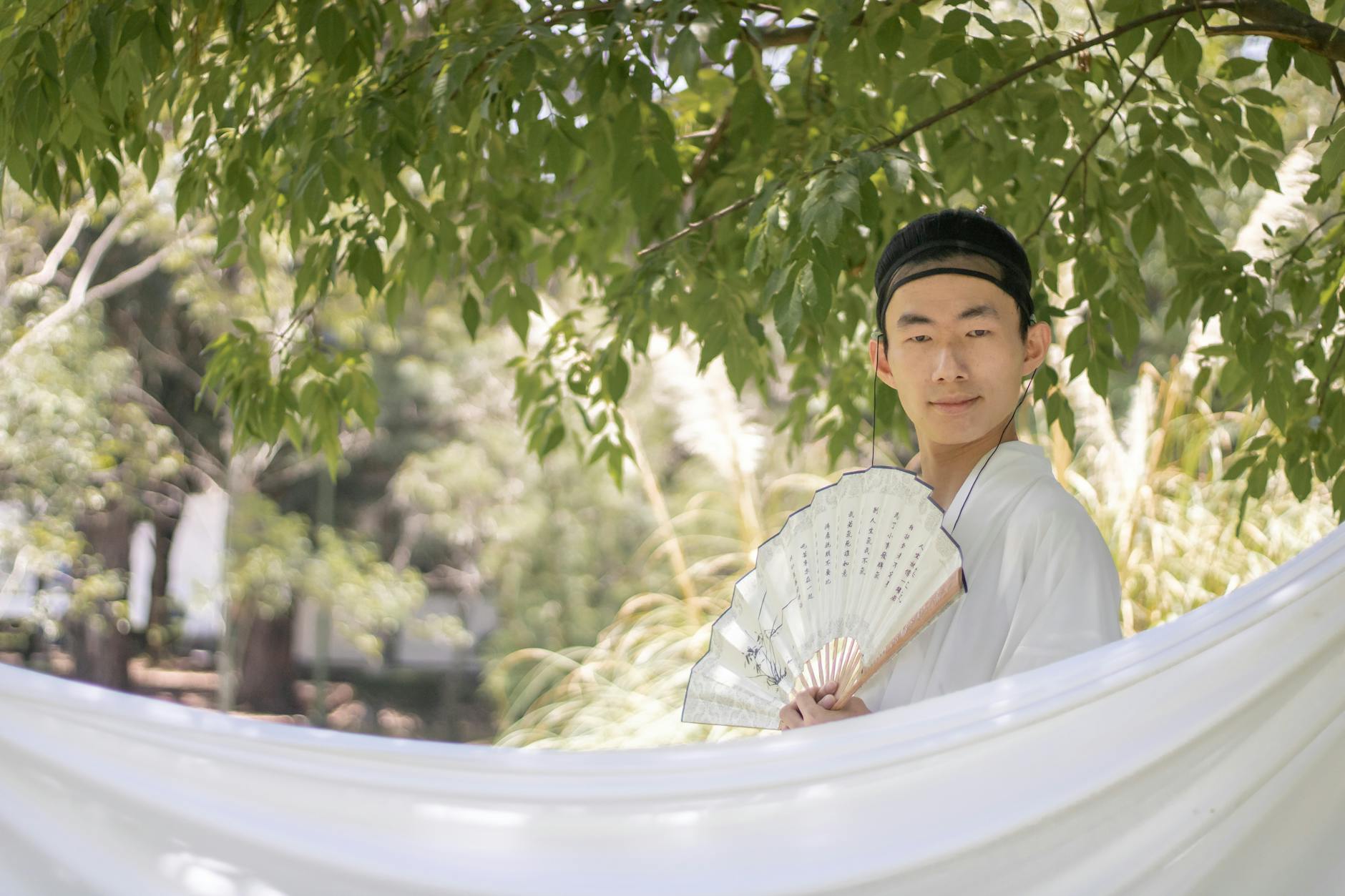 Mężczyzna w tradycyjnym azjatyckim stroju siedzi z wachlarzem pod drzewem